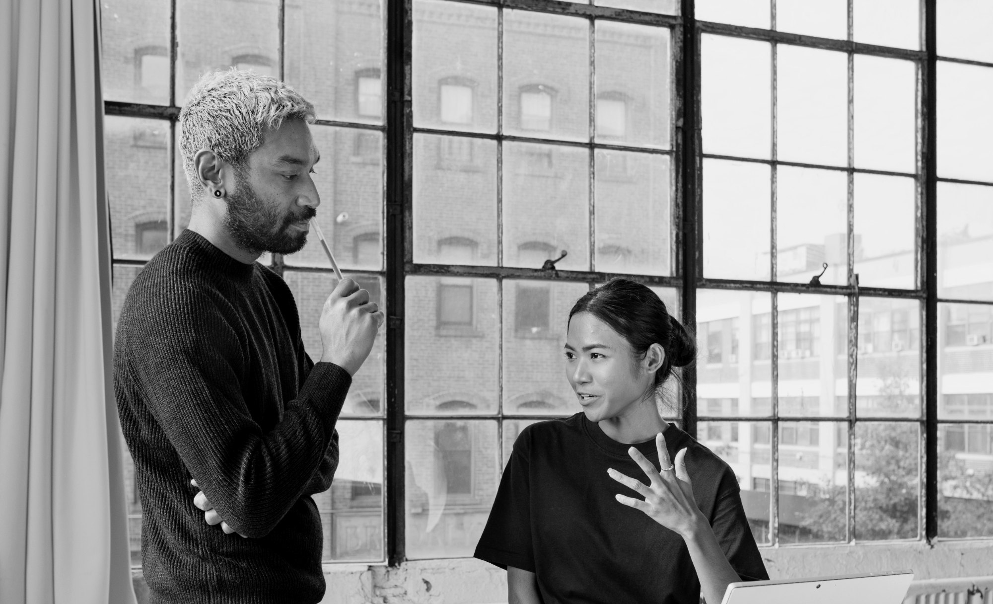 A man listens, arms folded, as a colleague explains by a tall warehouse window
